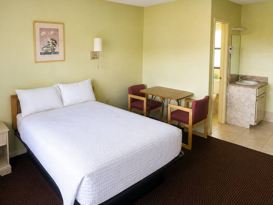 A hotel room with a bed, a desk, two chairs, a bedside table, a lamp, a mirror, and a bathroom with a sink and toilet.
