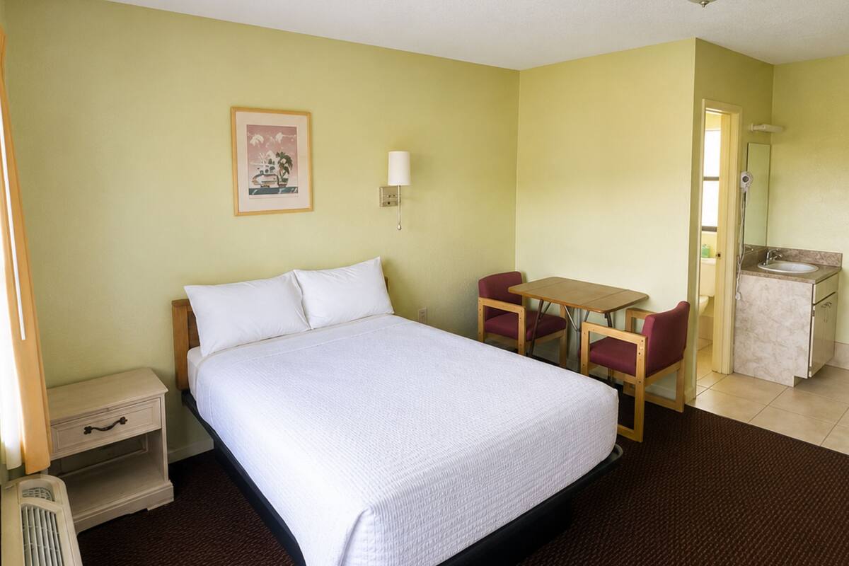 A hotel room with a bed, a desk, a chair, a small table, a nightstand, a lamp, a mirror, and a bathroom with a sink and toilet.