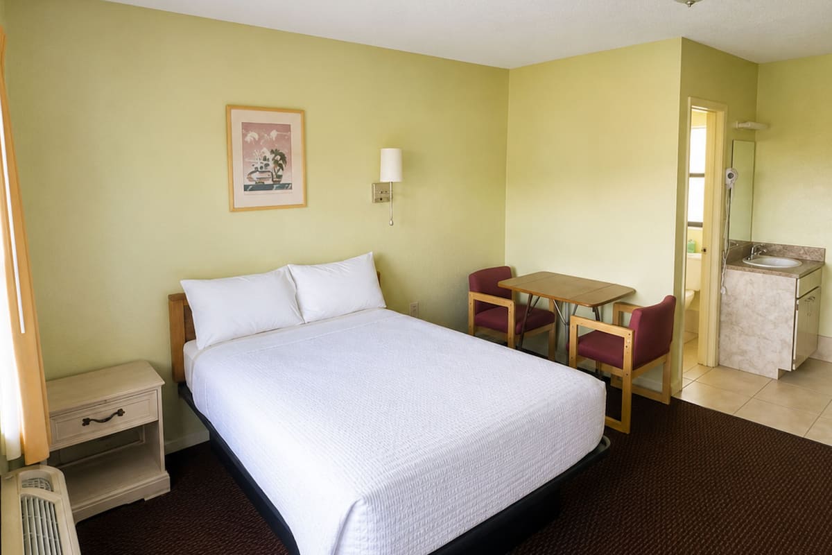 Ein Hotelzimmer mit Bett, Schreibtisch, Stuhl, einem kleinen Tisch, Nachttisch, Lampe, Spiegel und einem Badezimmer mit Waschbecken und Toilette.