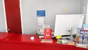 A hotel reception desk with a red counter, a desktop computer, a mouse, and various travel brochures.