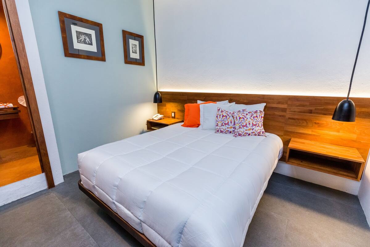 A neatly made bed with a white comforter and a wooden headboard. There are two bedside tables, one with a lamp and the other with a phone. The room has light blue walls and a wooden floor.
