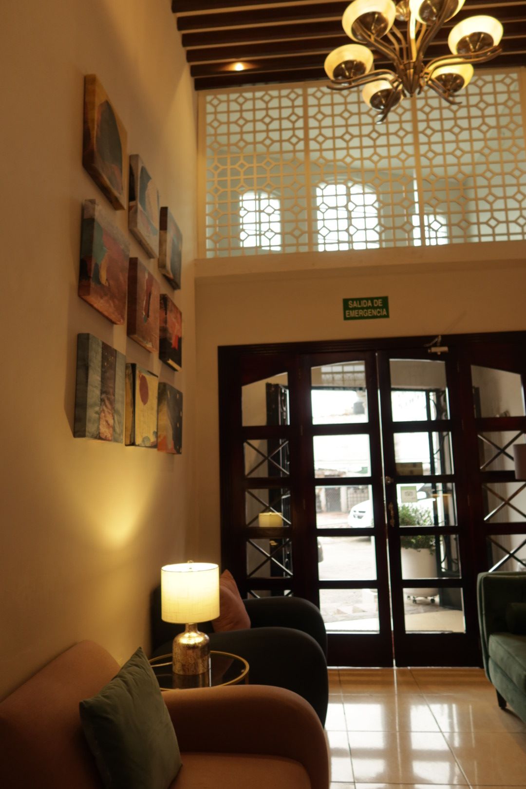 A reception area with a sofa, a lamp, artwork on the wall, a sign indicating an emergency exit, and a view through a glass door.