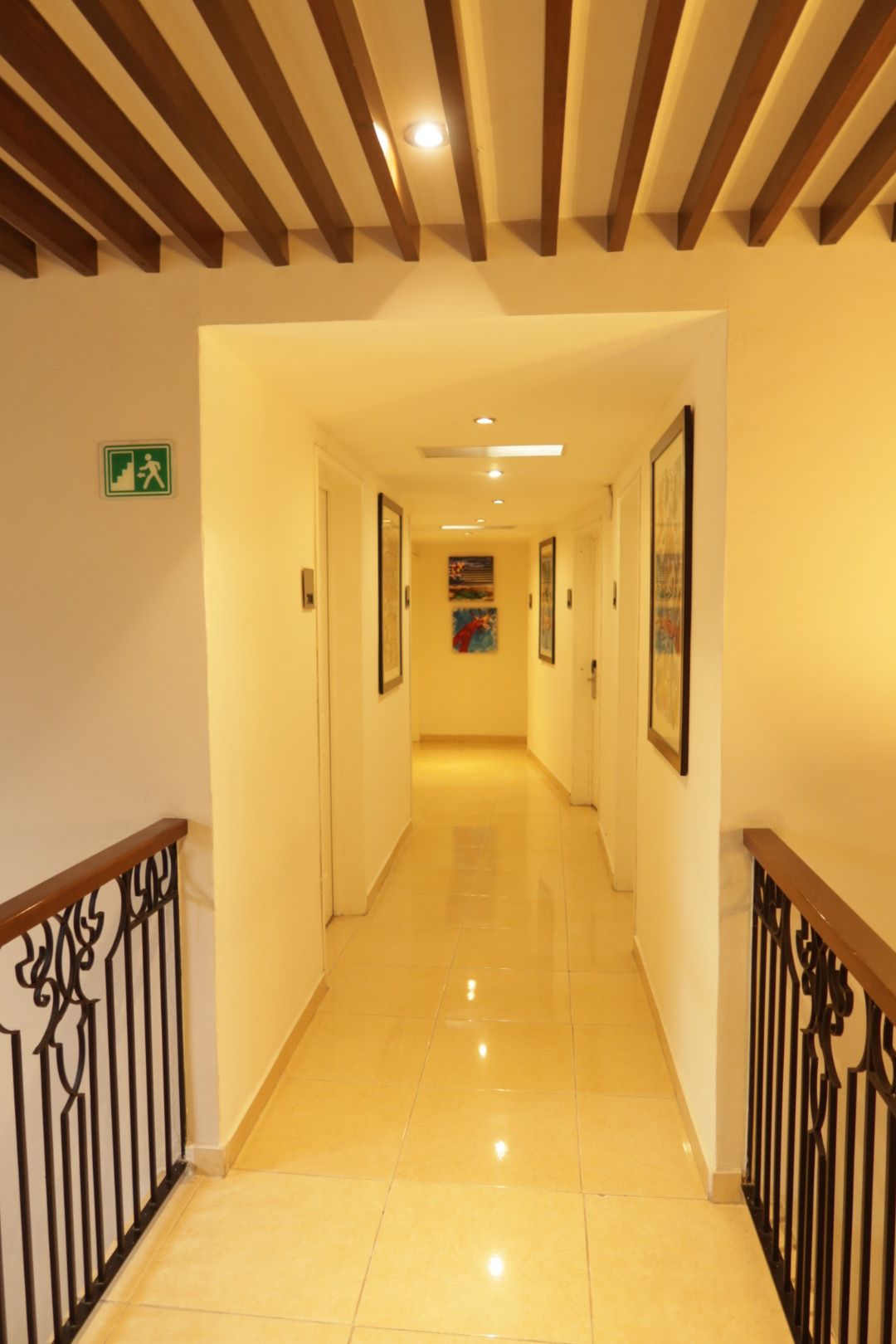 A hotel corridor with a polished floor, wooden ceiling beams, and a green emergency exit sign.