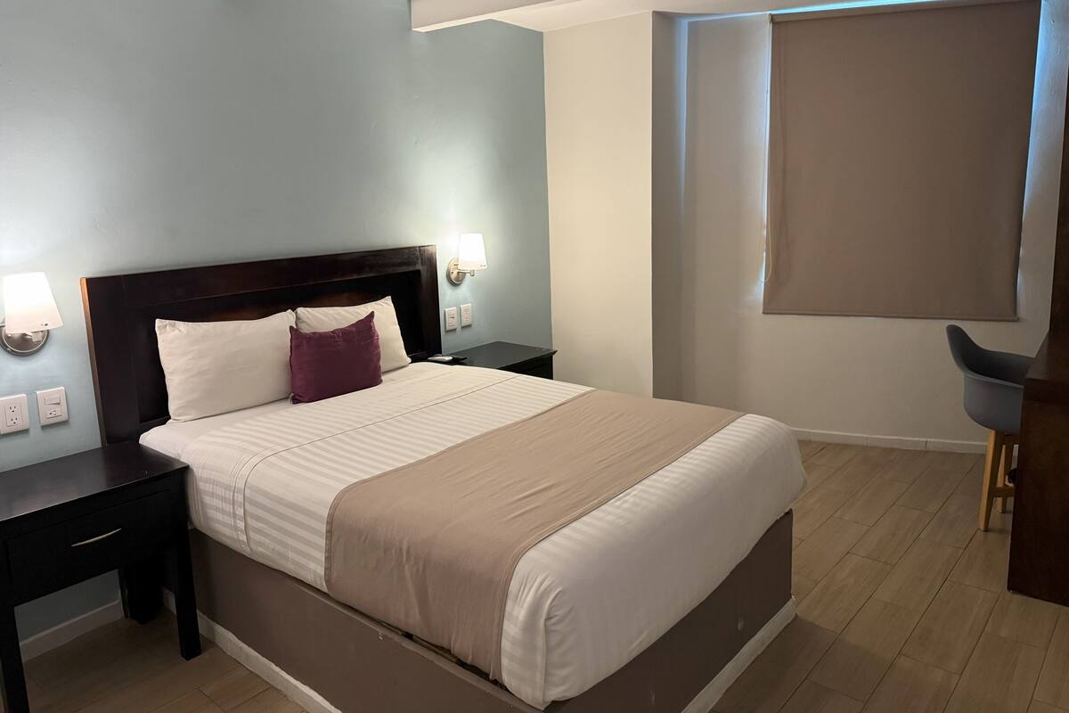 A neatly made bed with a headboard, two bedside tables with lamps, a wooden floor, and a large window with a blind.