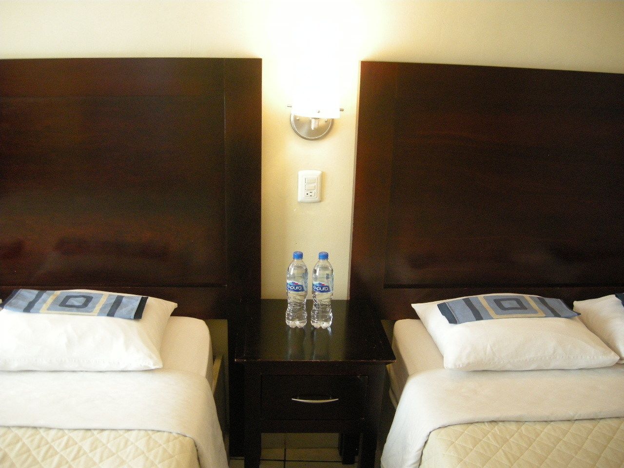A hotel room with two beds, each with a pillow and a blanket. There is a small table between the beds with two bottles of water and a lamp on it.