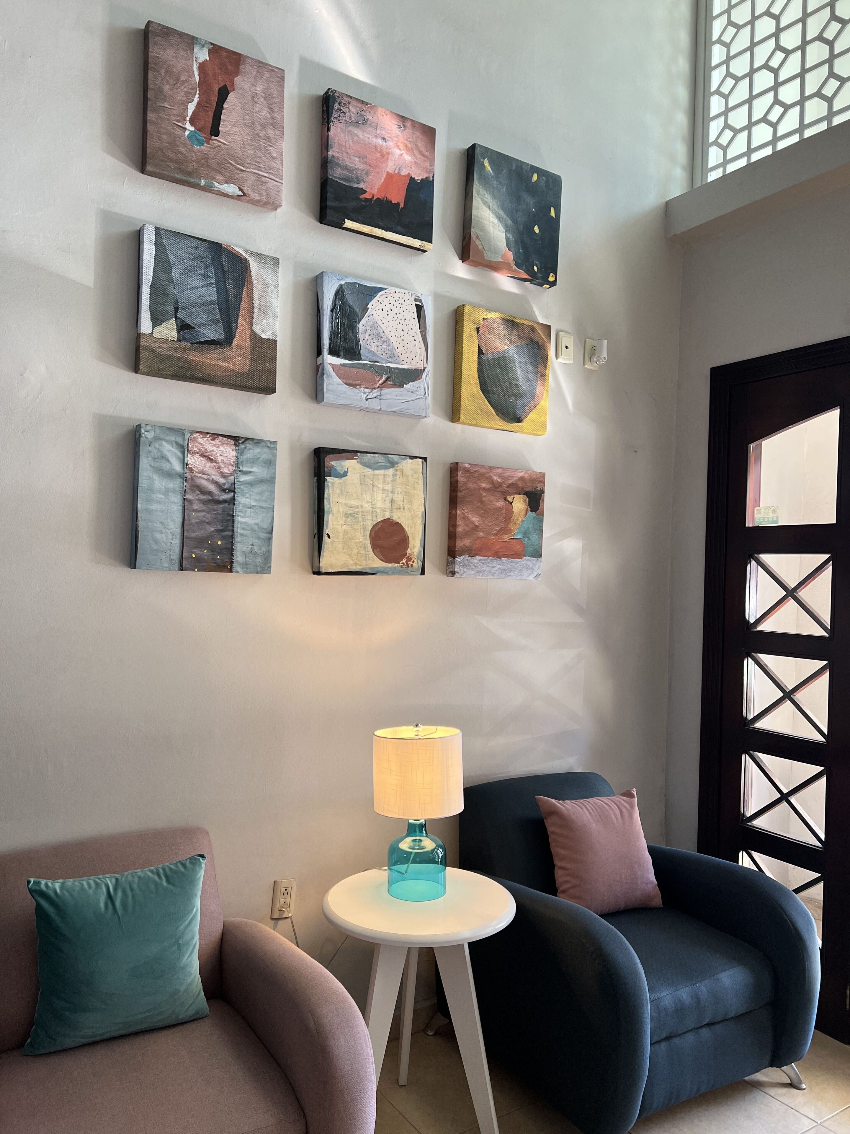 A room with a seating area featuring two armchairs and a small table with a lamp. There are several abstract paintings on the wall.