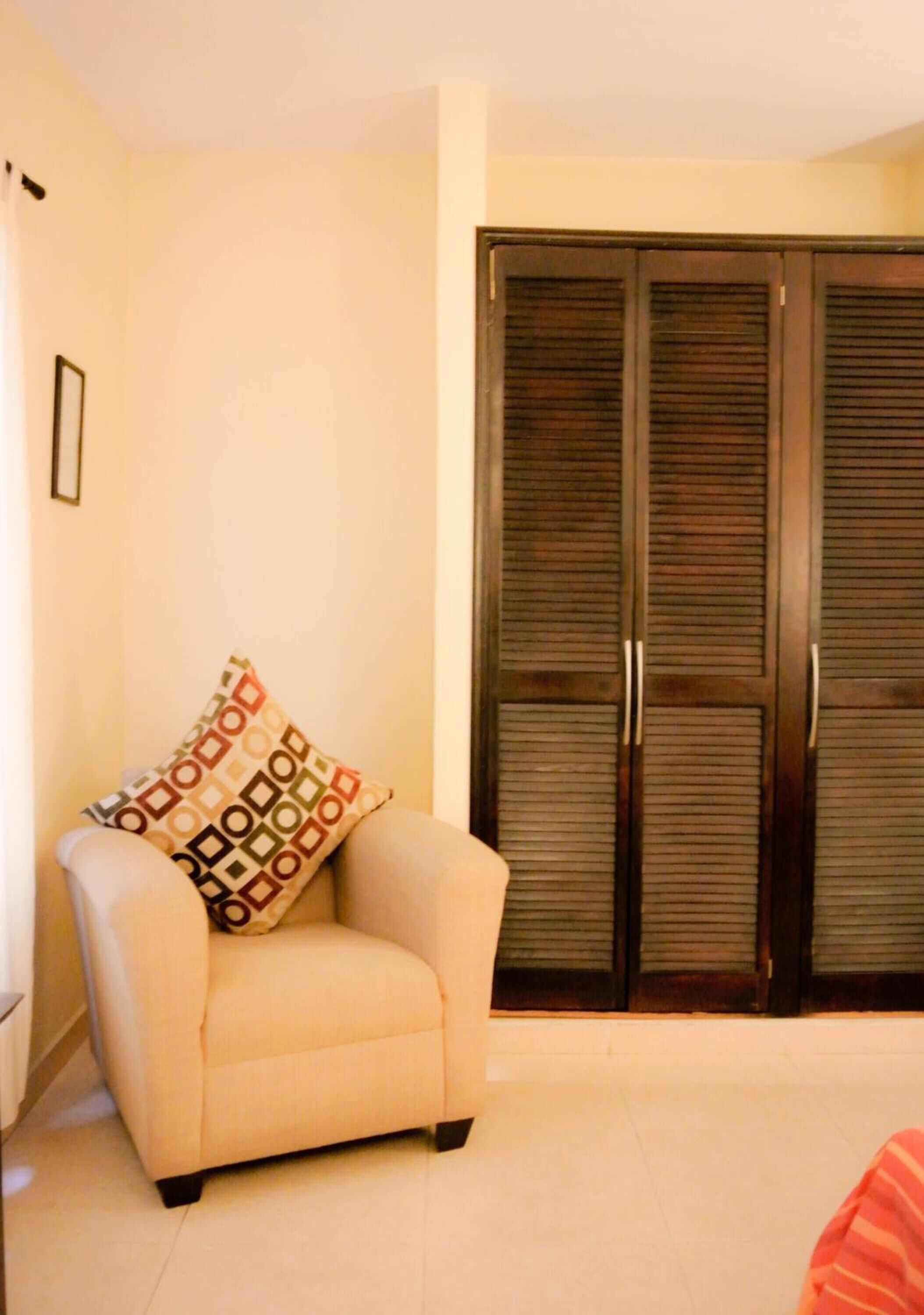 A room with a beige armchair, a patterned pillow, and a large dark wooden wardrobe.