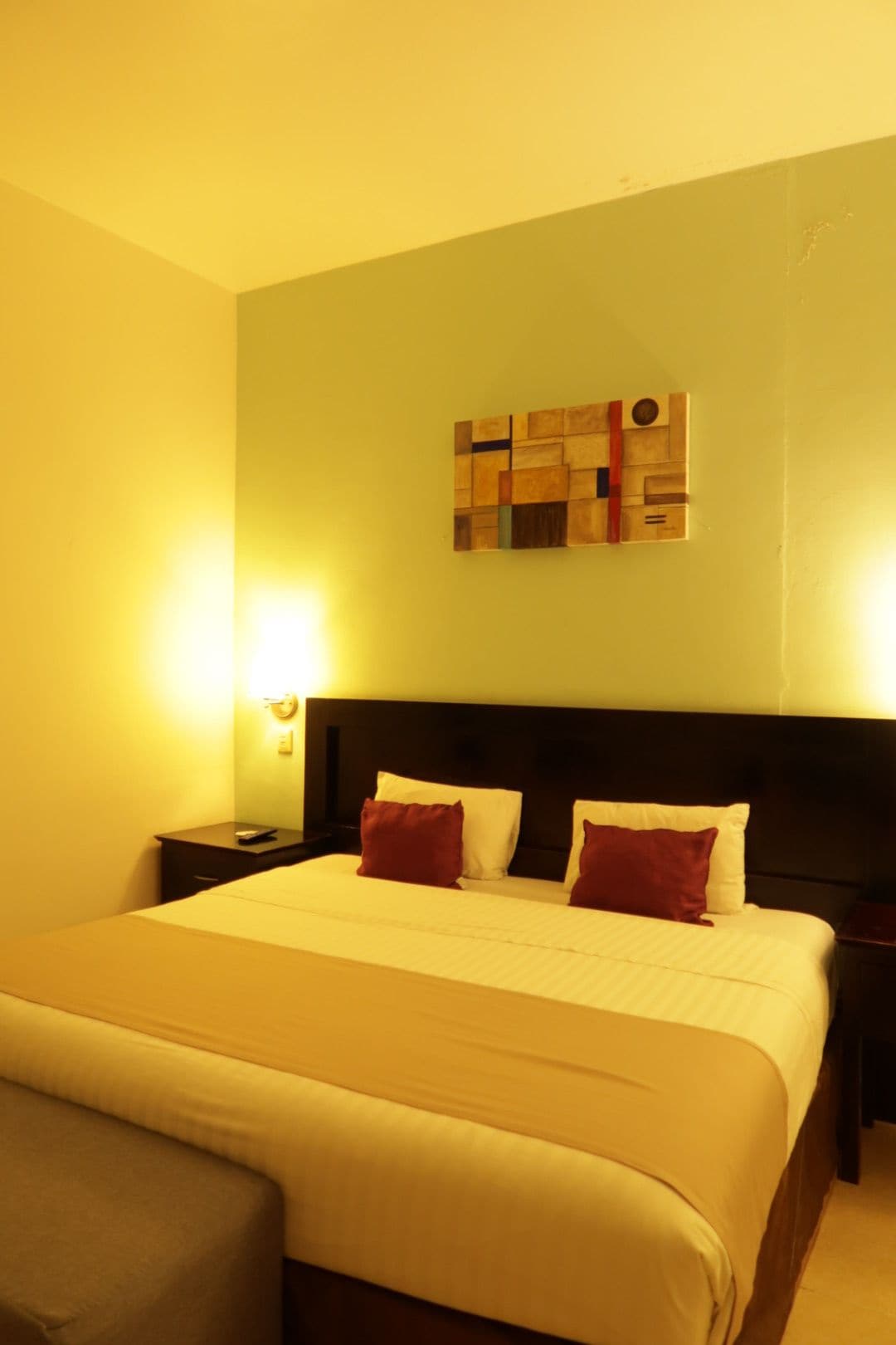 Une chambre d’hôtel avec un grand lit, deux tables de chevet, une petite table d’appoint, une œuvre d’art accrochée au mur et une lampe murale.