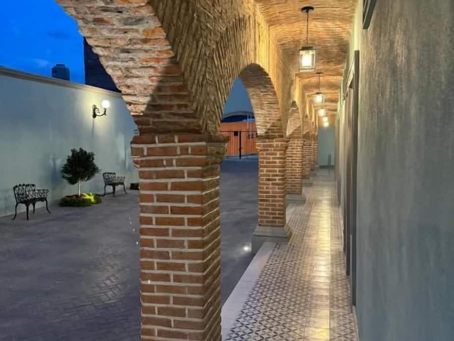 A corridor with brick arches and patterned flooring, leading to a building with a flat roof.