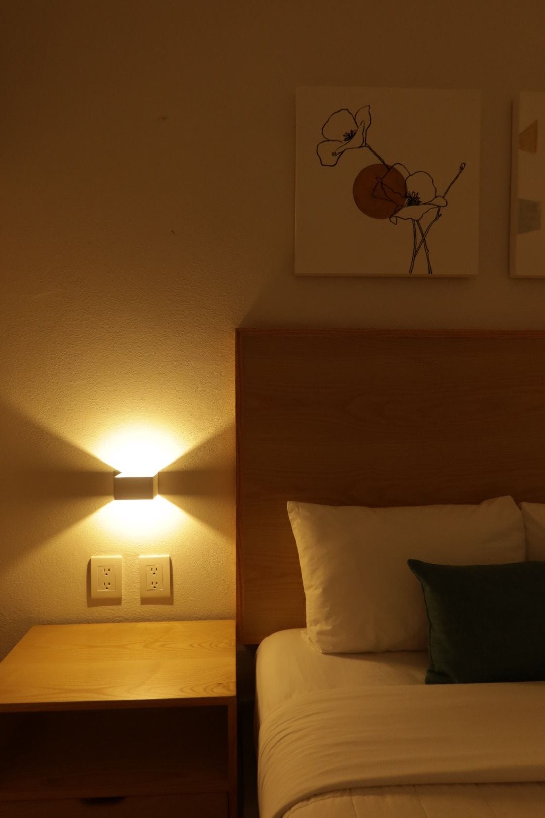 A hotel room with a bed, a bedside table, a wall-mounted light, and a painting.