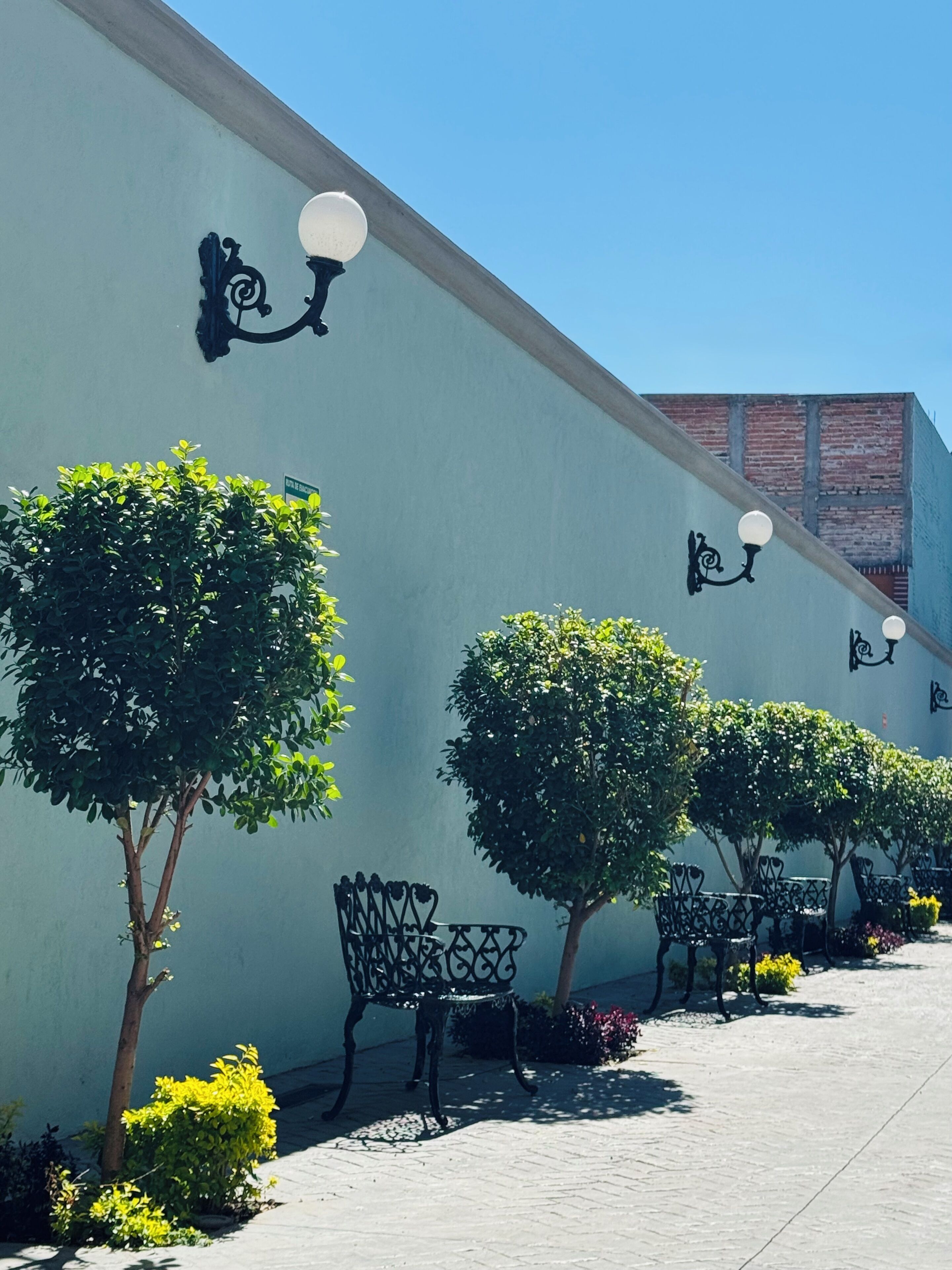A pathway lined with trees and benches, with wall-mounted lights and a clear blue sky.