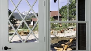A view through a window with a white frame, revealing a patio area with wooden chairs and a table, a garden with potted plants, and a mountain range in the background. - River Rock Cottages (Estes Park)