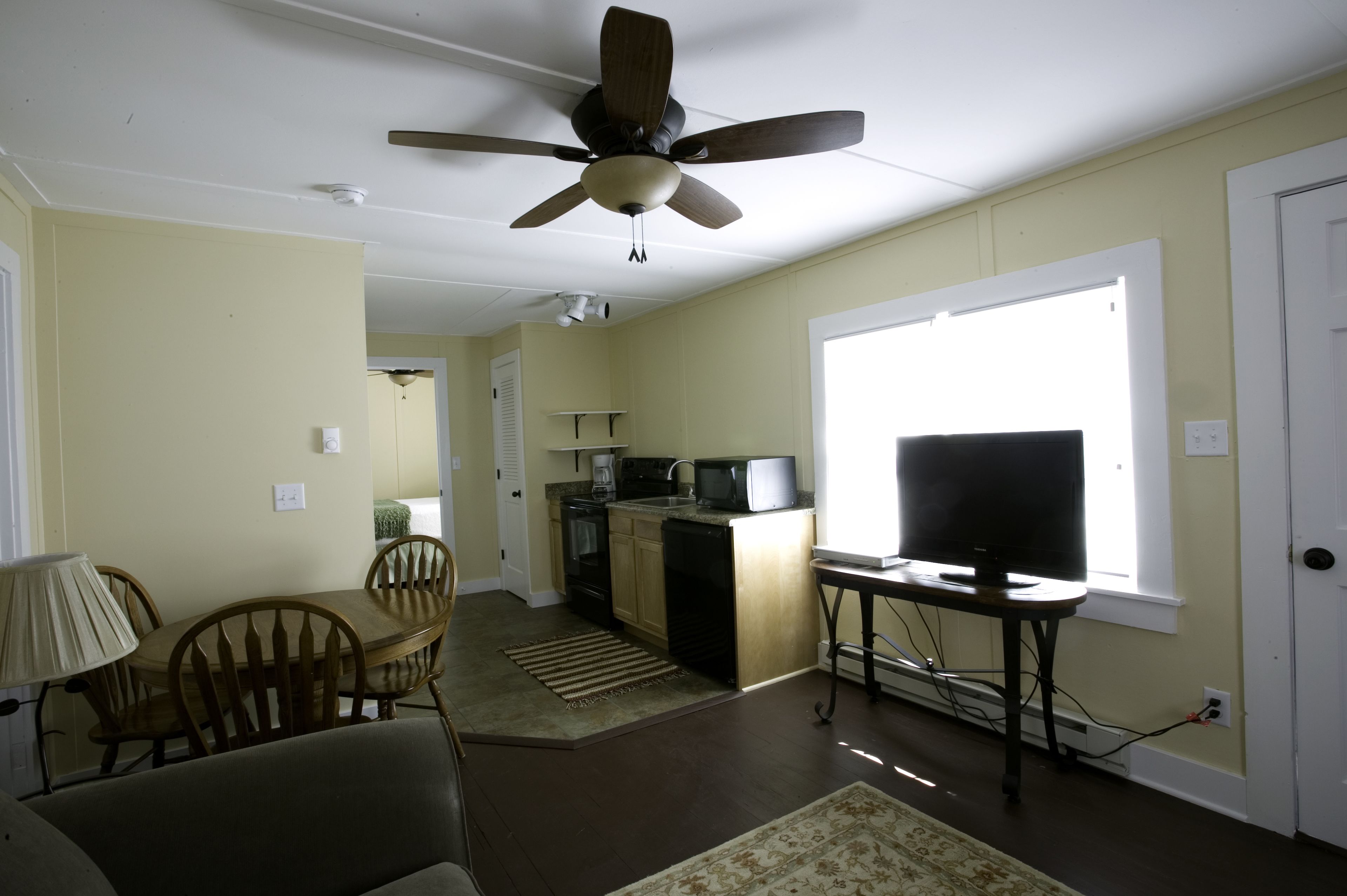 Un salon doté d’un ventilateur de plafond, une table à manger avec des chaises, un coin cuisine avec un four à micro-ondes et une grande fenêtre.