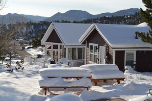Eine Hütte mit verschneitem Deck und Bänken, umgeben von verschneiter Landschaft und Bergen.