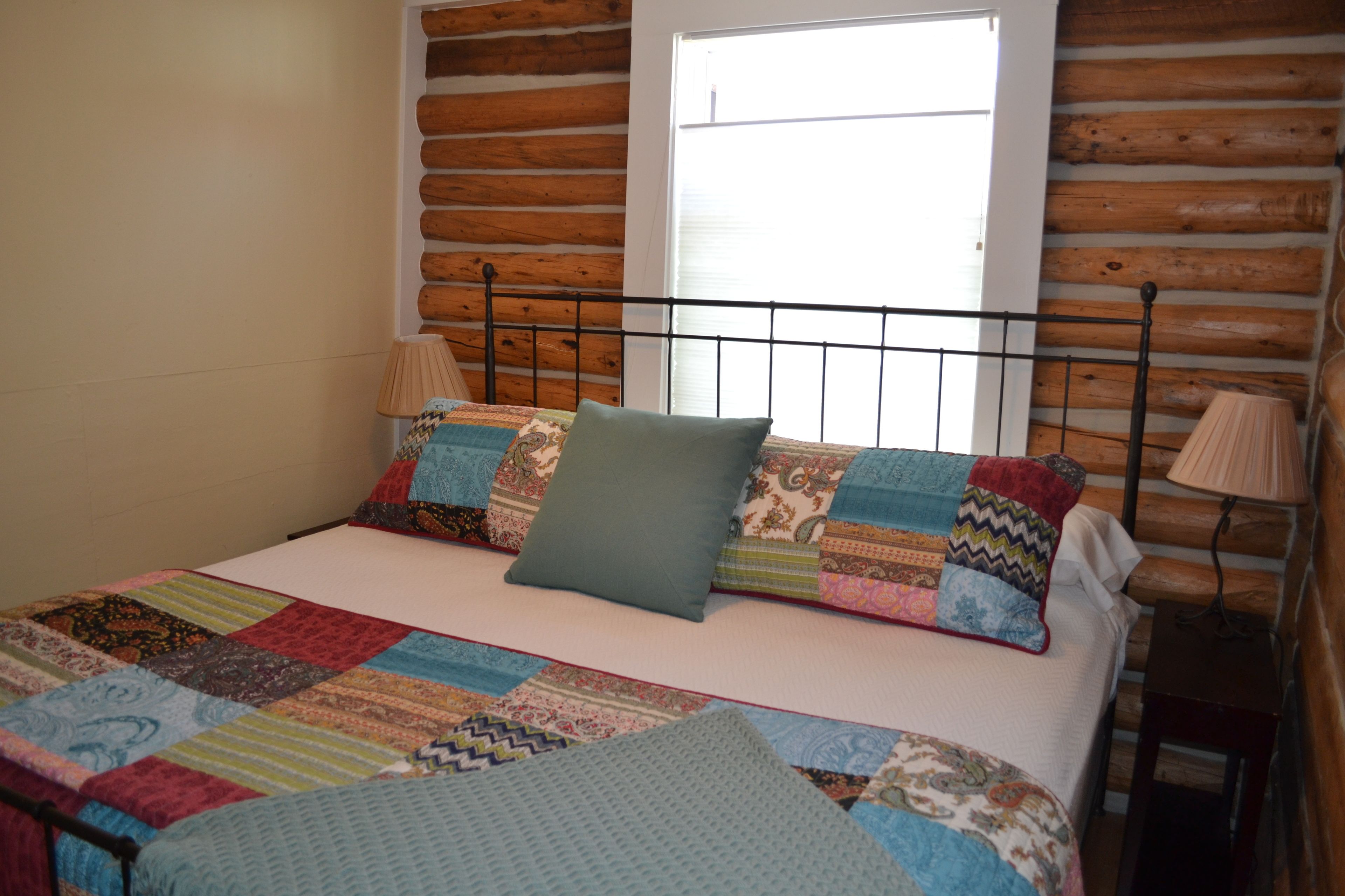 A bed with a colorful quilt and pillows, a wooden log wall, and a window with blinds.