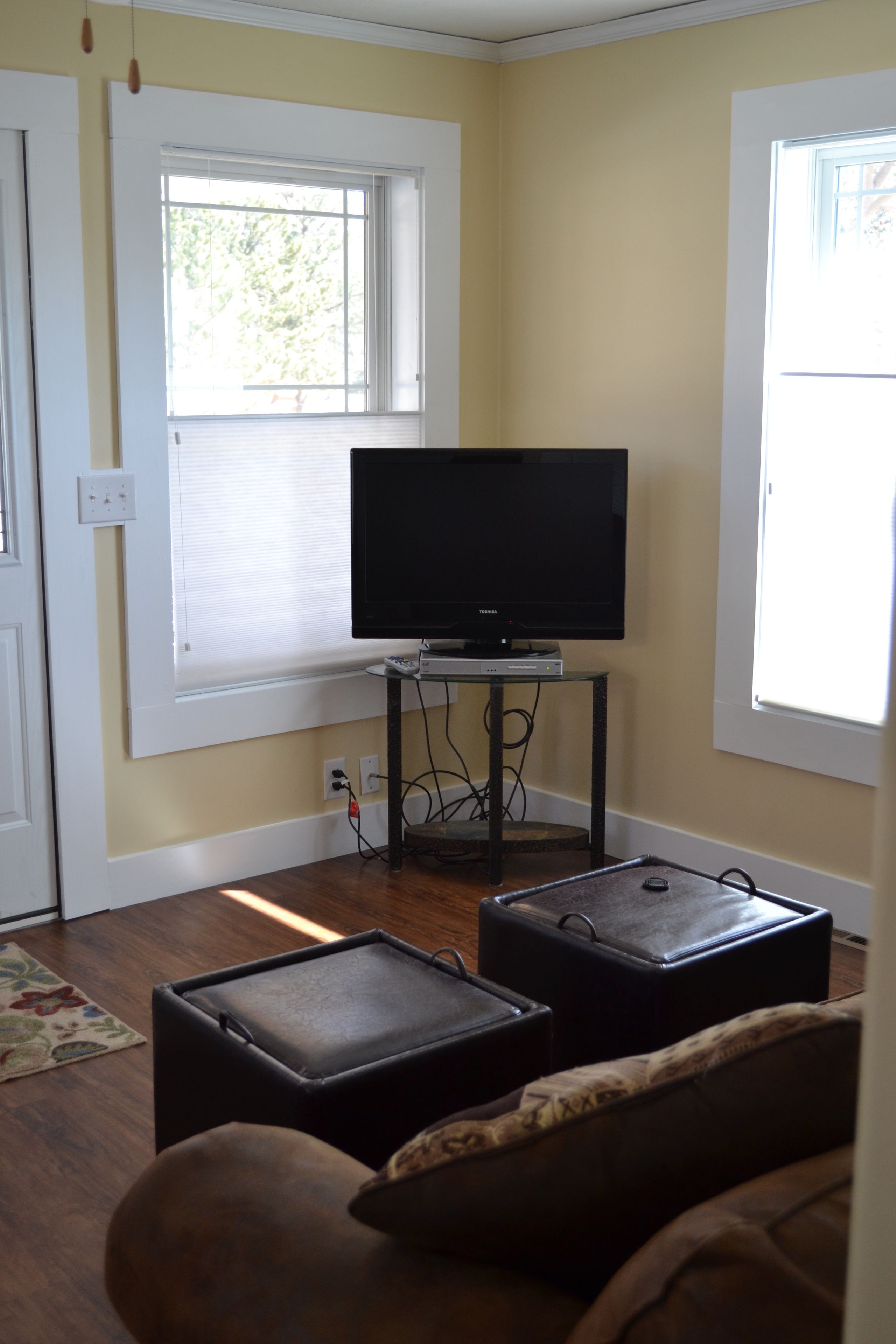 A living room with a television on a stand, two ottomans, a sofa, and a window with blinds.