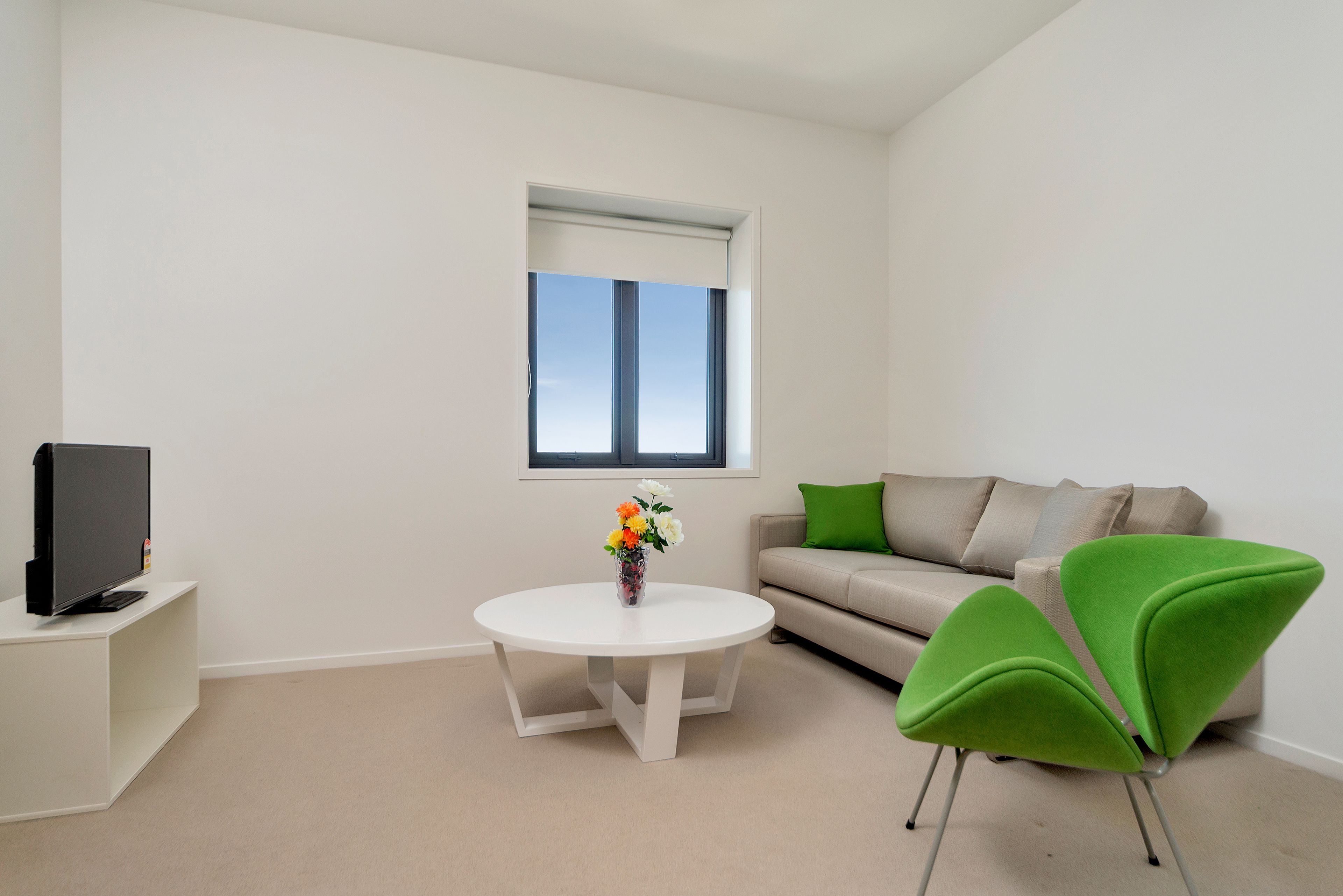 A modern living room with a white round table, a green armchair, a beige sofa, and a flat-screen TV on a white stand.