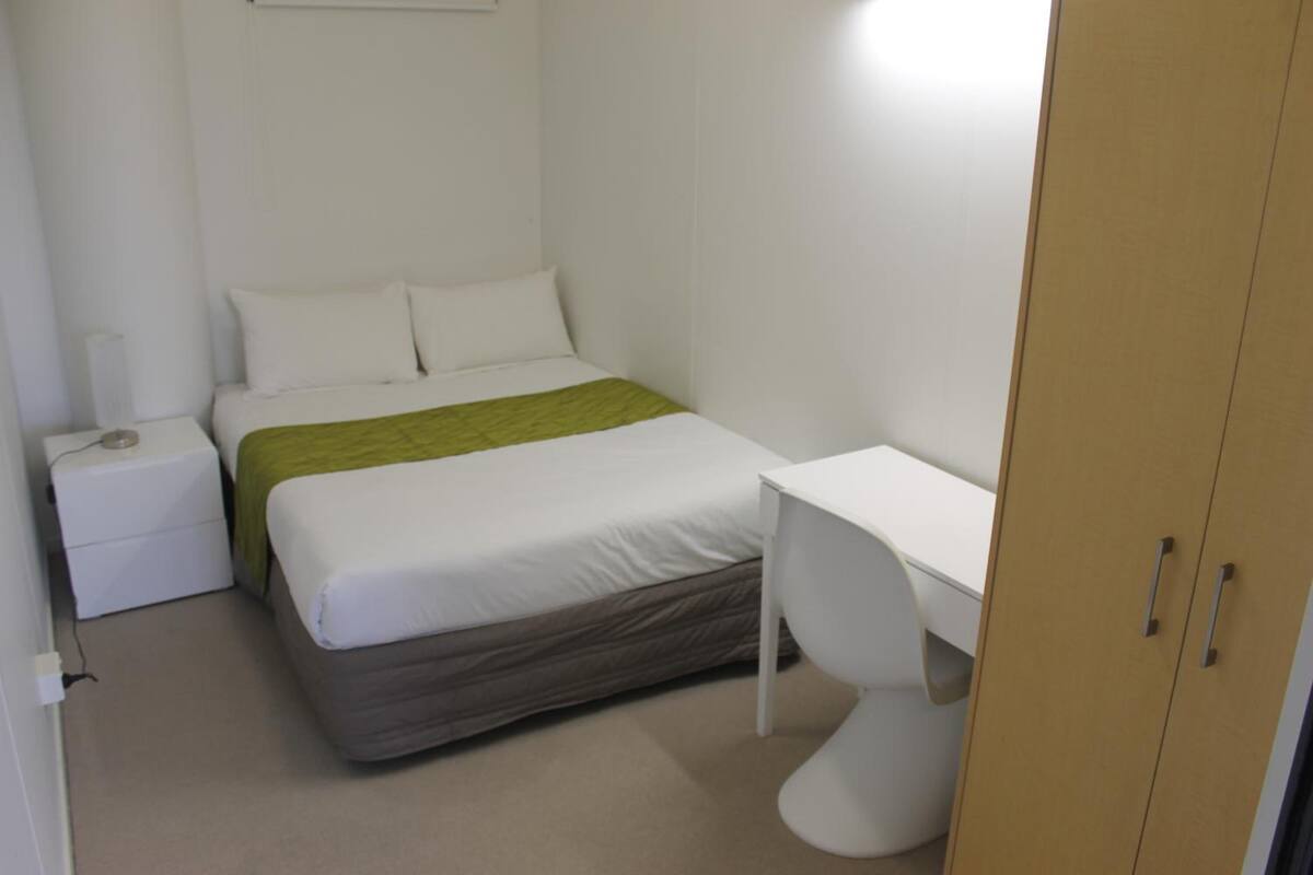 A small, clean room with a bed, a bedside table, a desk, and a wardrobe.