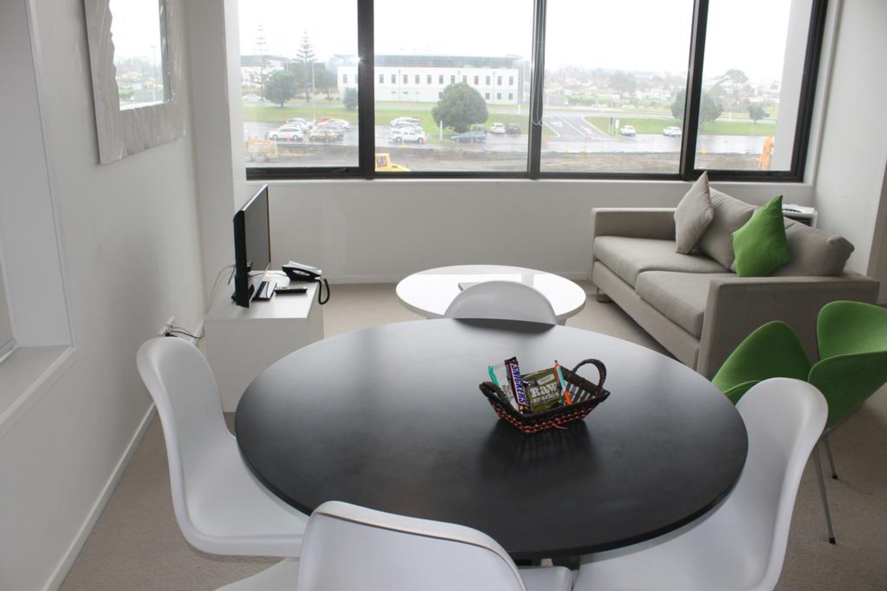 A modern office space with a round table, white chairs, a sofa, and a view of a building through large windows.