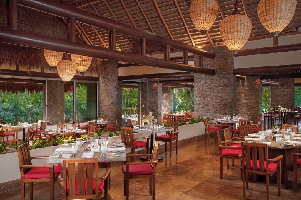 A spacious restaurant with multiple dining tables and chairs, large windows offering views of greenery, and hanging lantern-style lights.