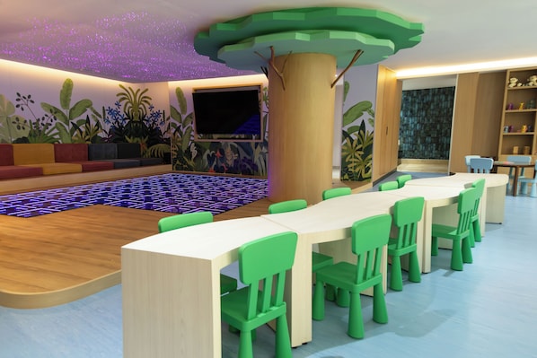 Un área de juegos infantiles con una mesa grande y sillas, un televisor de pantalla plana y un mural colorido en la pared.