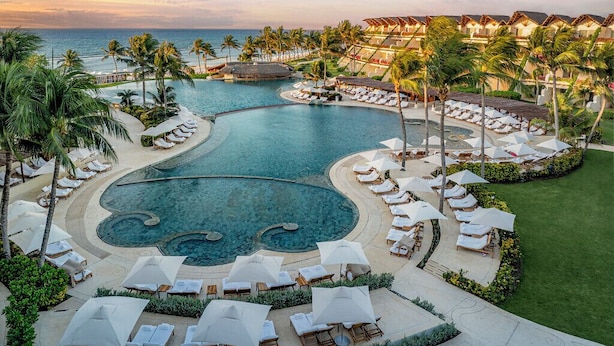 A resort pool area with multiple sun loungers and umbrellas, surrounded by palm trees and a view of the ocean. - Ambassador at Grand Velas All Inclusive (Playa del Carmen)