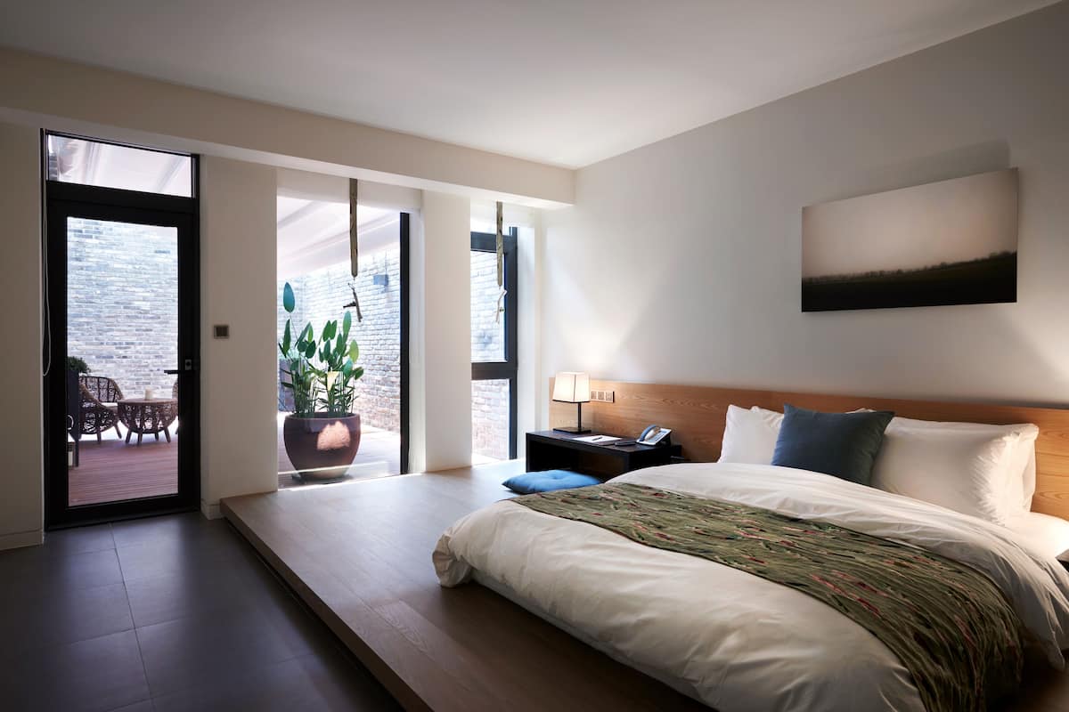 Quarto de hotel moderno com cama grande, mesa de cabeceira, planta em vaso e vista para o exterior por uma porta deslizante.