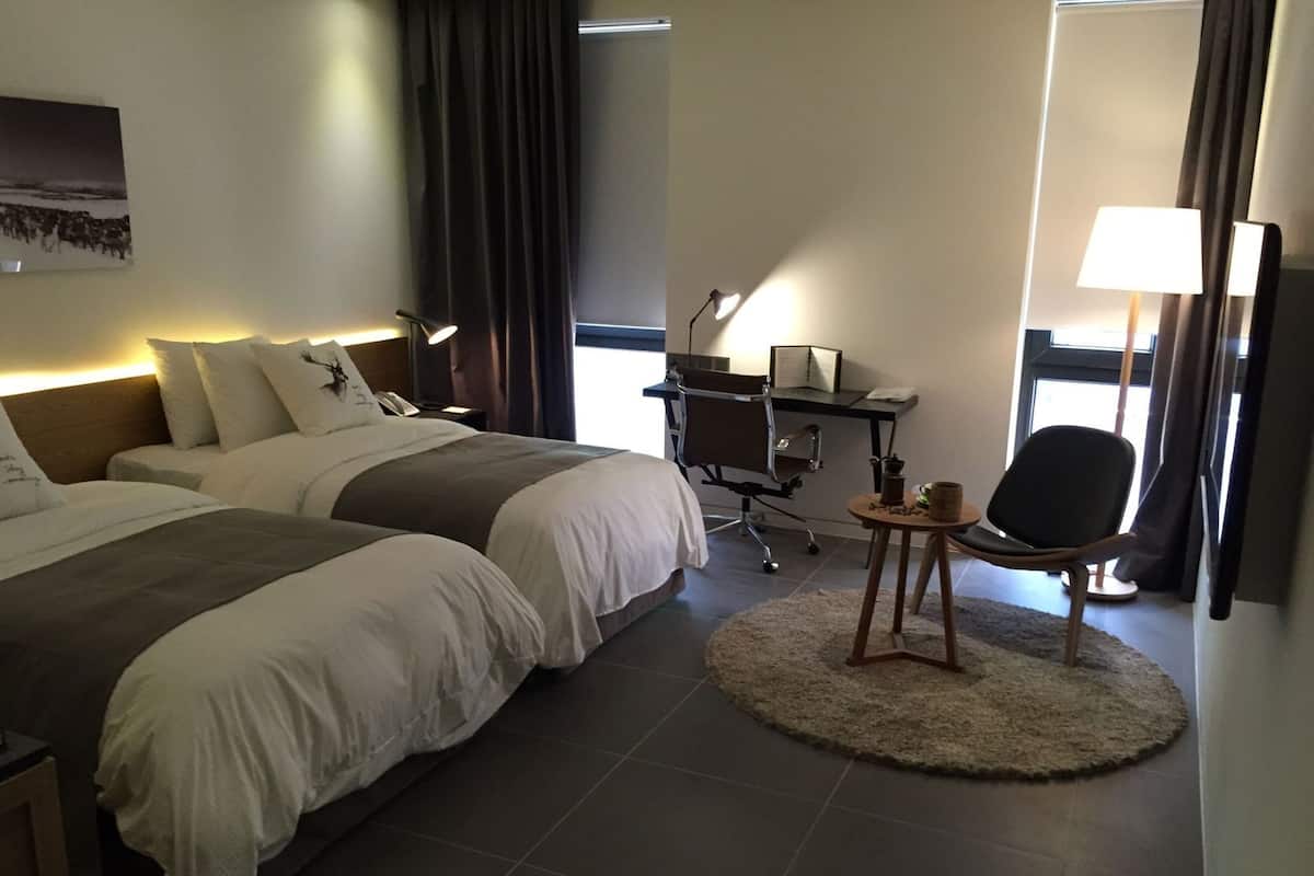 Quarto de hotel com duas camas, uma escrivaninha com cadeira, uma mesinha de cabeceira com abajur e uma janela grande.