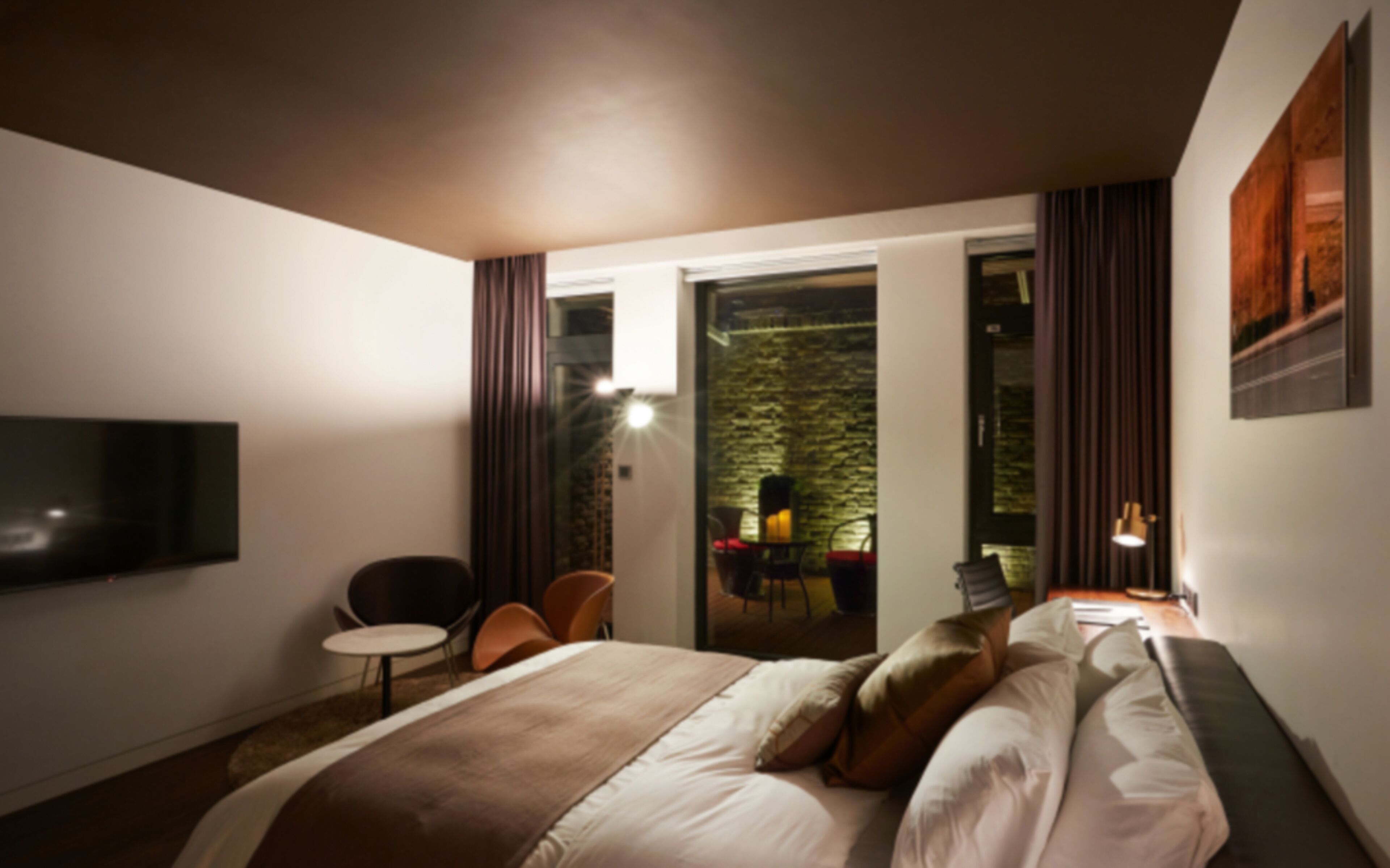 A hotel room with a bed, a TV, a small table, and a chair. There is a balcony with a table and chairs outside the room.