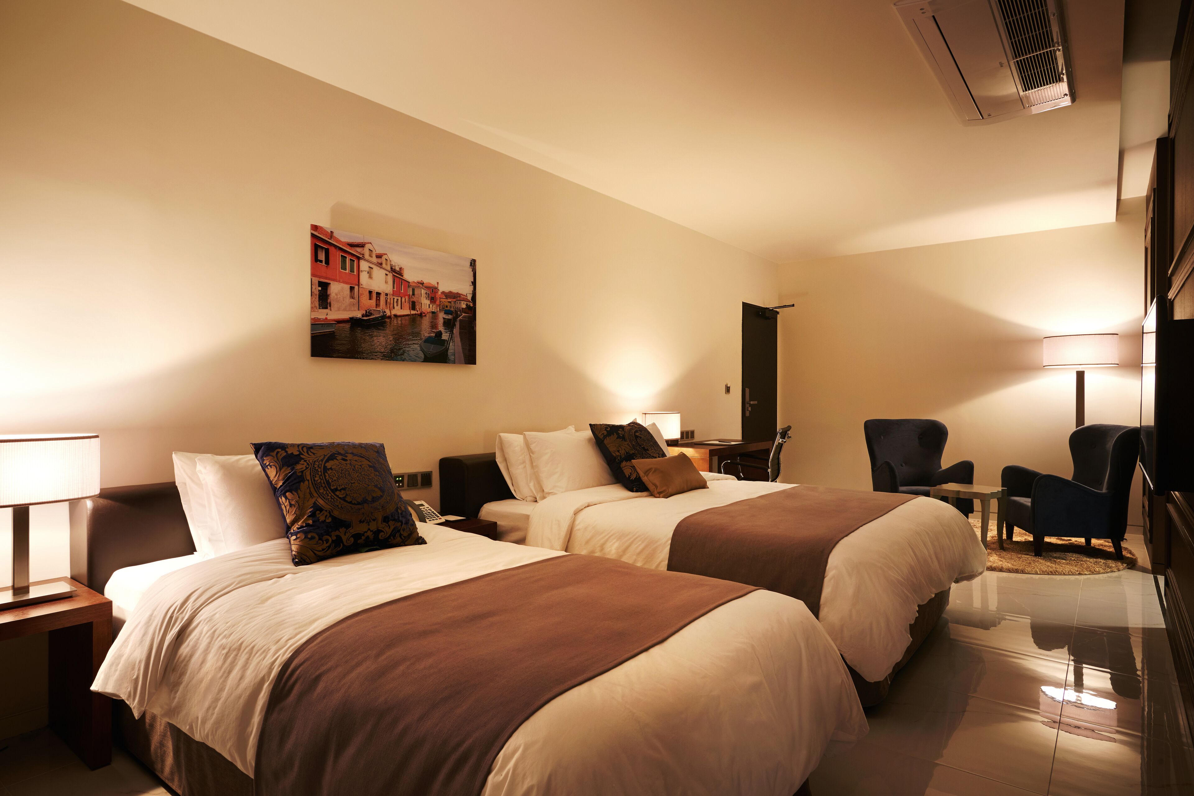 A hotel room with two beds, a desk, a chair, and a painting on the wall.