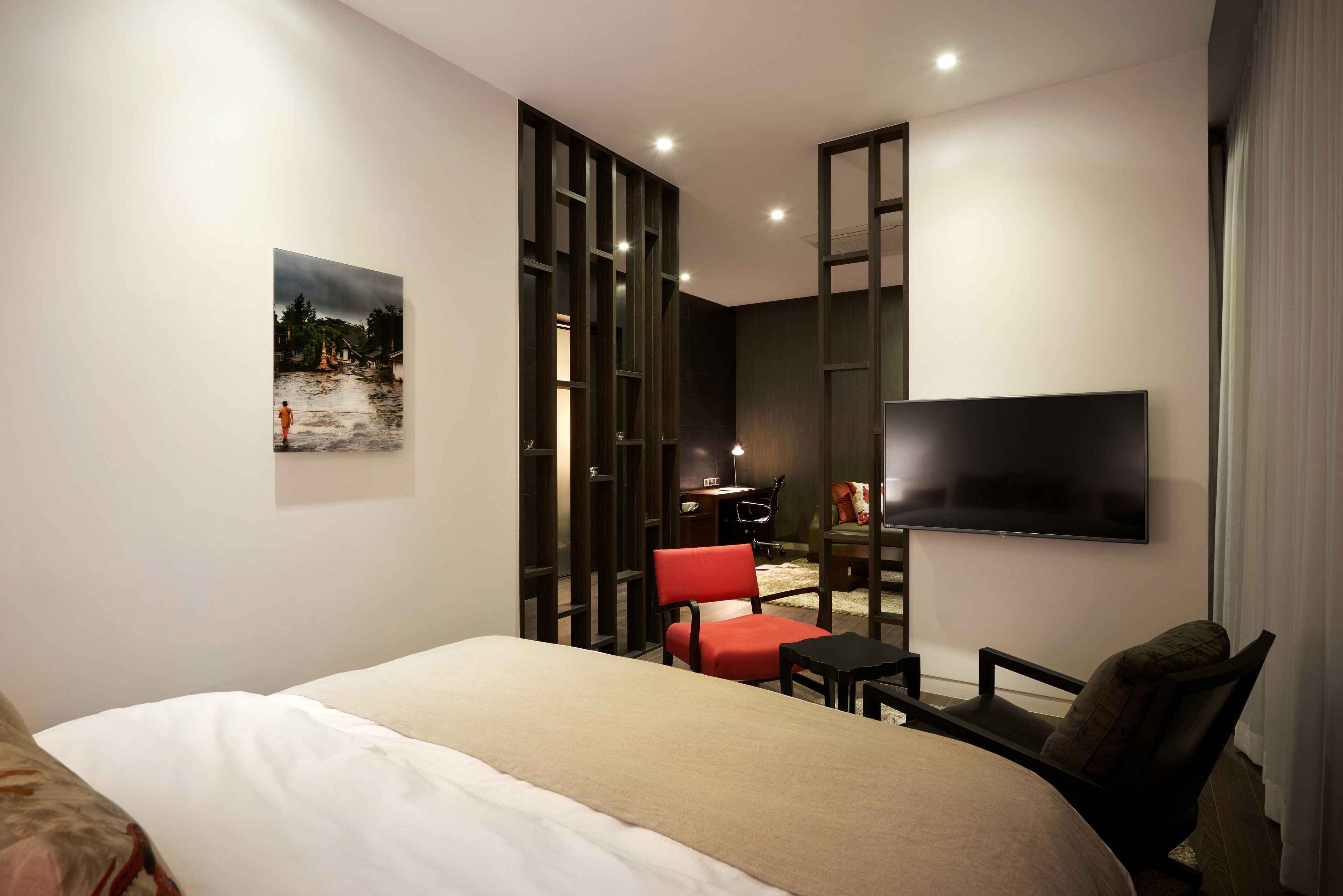 A modern hotel room with a large bed, a flat-screen TV mounted on the wall, a desk with a chair, and a red armchair.