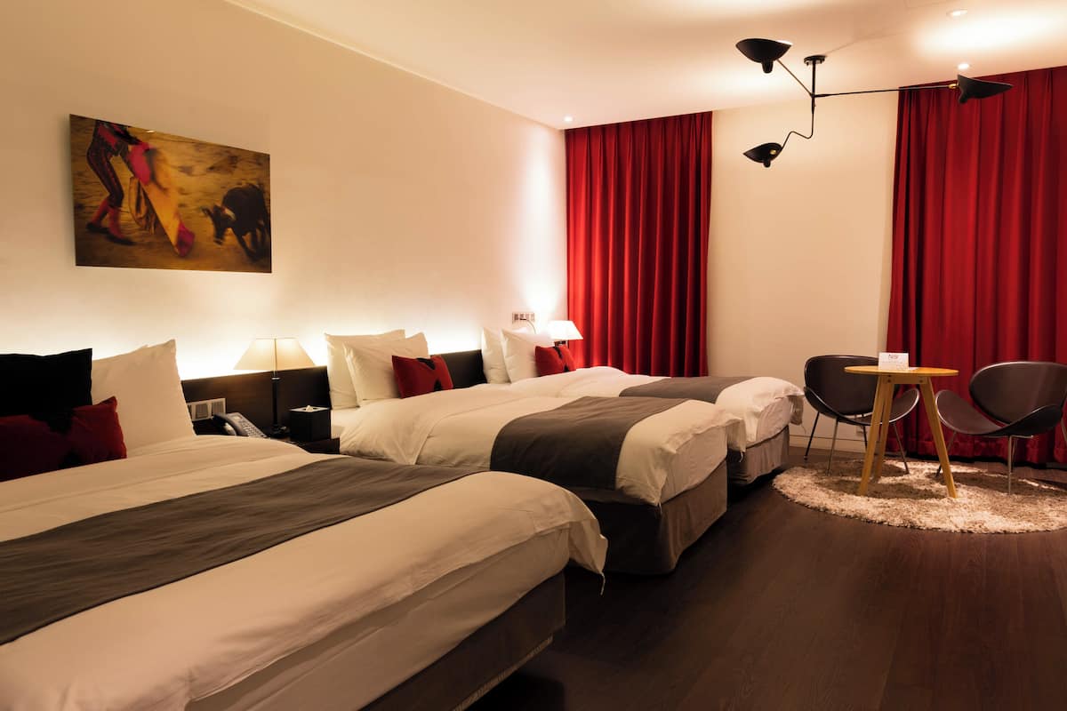 Quarto de hotel com duas camas, uma escrivaninha, cadeiras, cortina vermelha e um quadro na parede.