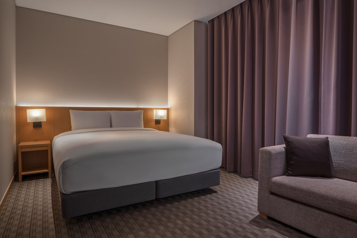 A hotel room with a large bed, a nightstand with a lamp, a sofa, and a curtain.