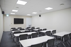 A conference room with white tables and black chairs, a large flat-screen TV on the wall, and a door with a light switch.