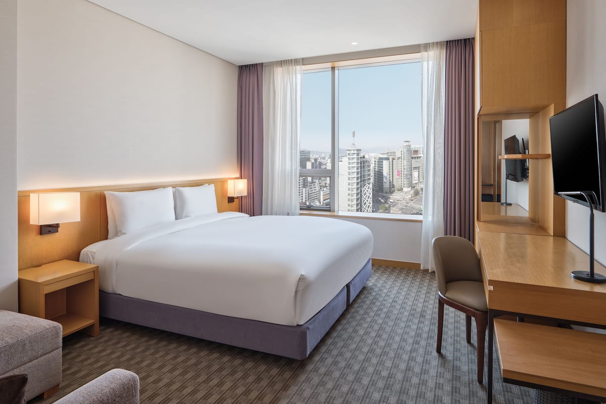 A hotel room with a large bed, a desk with a chair, a TV, and a city view through the window.