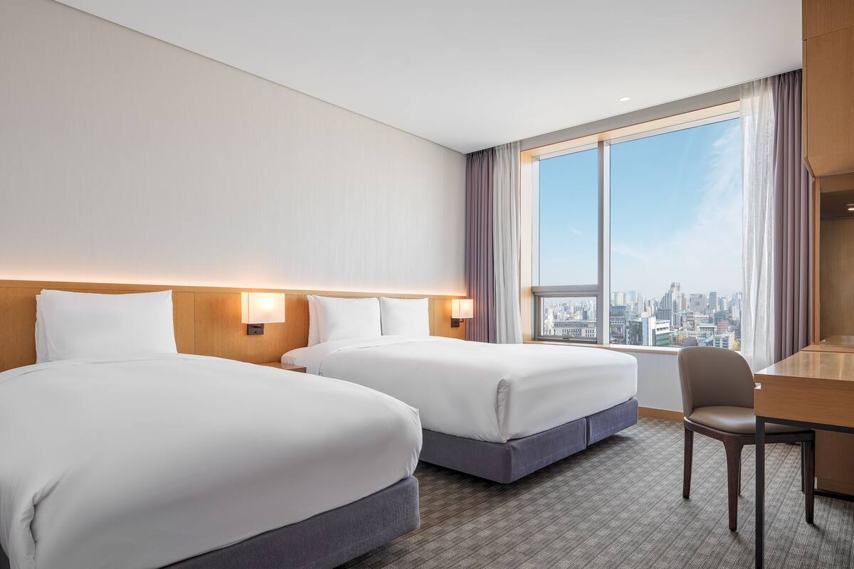 A hotel room with two beds, a desk, and a chair. The room has a city view through large windows.