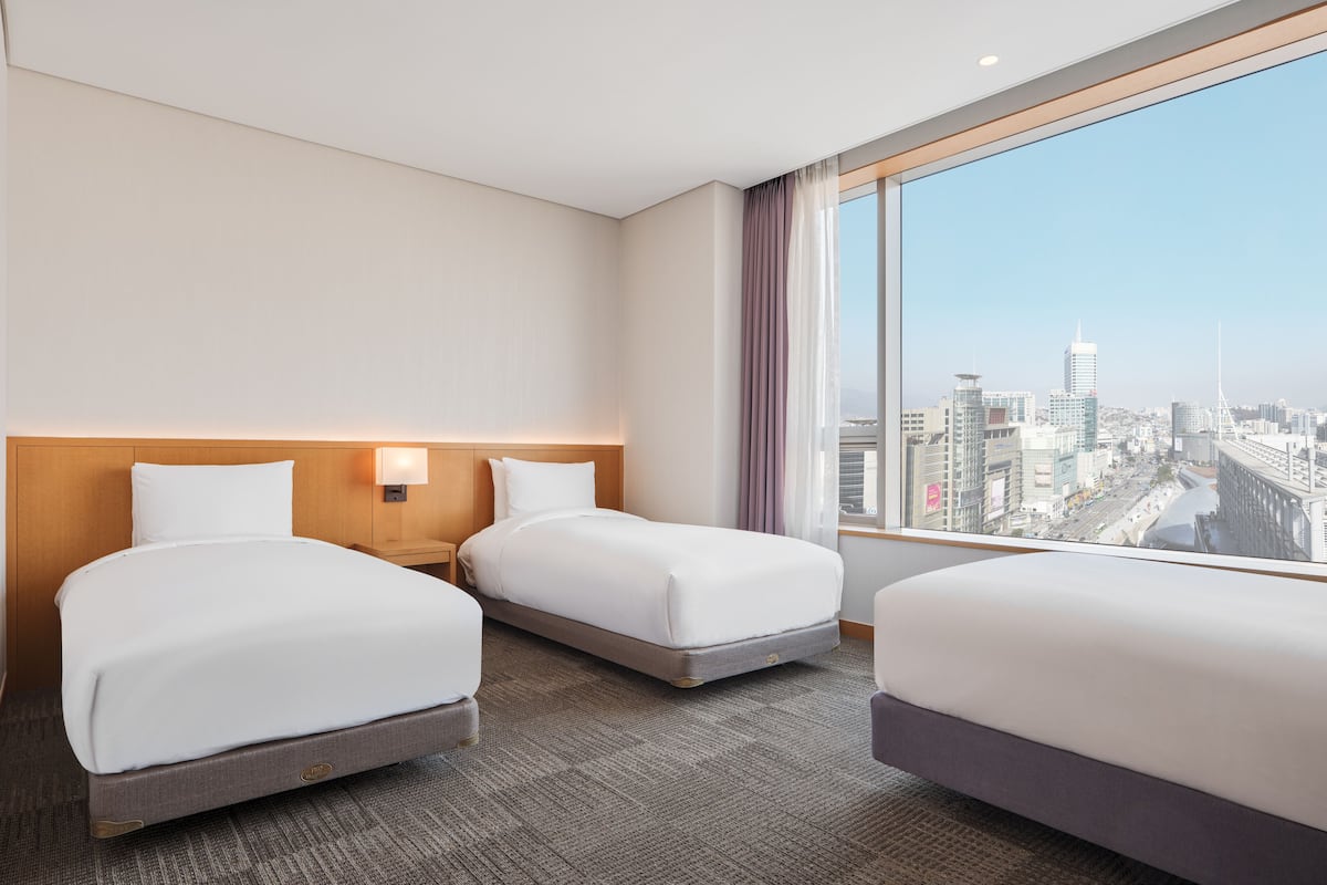 A hotel room with two beds, a nightstand, a large window with a city view, and a wooden headboard.