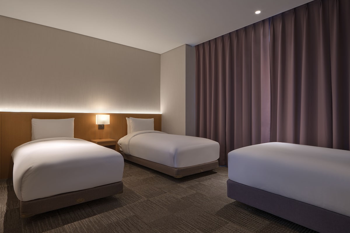 A hotel room with two beds, a nightstand, a wall-mounted lamp, and a large window with curtains.