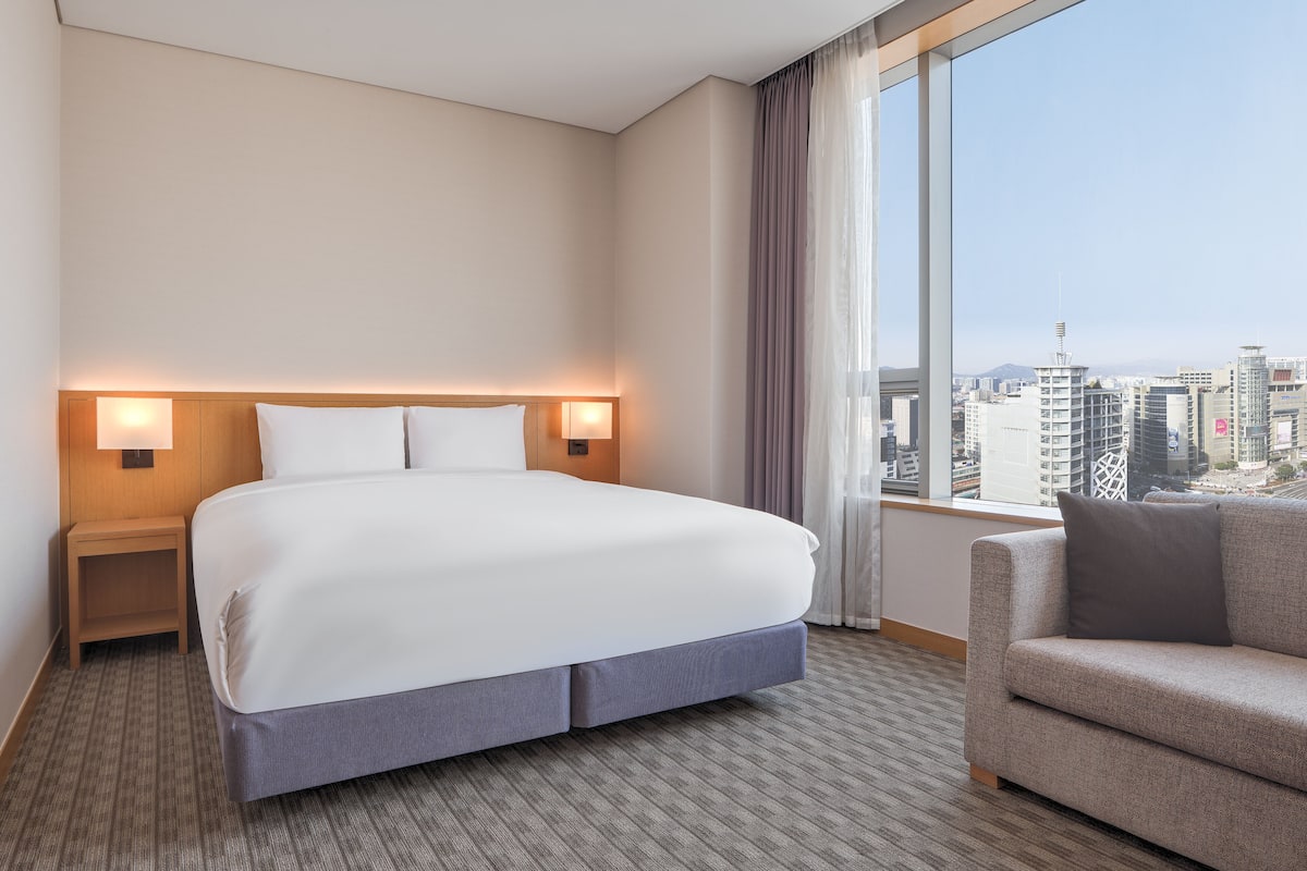 A hotel room with a large bed, two bedside lamps, a view of the cityscape, and a sofa.