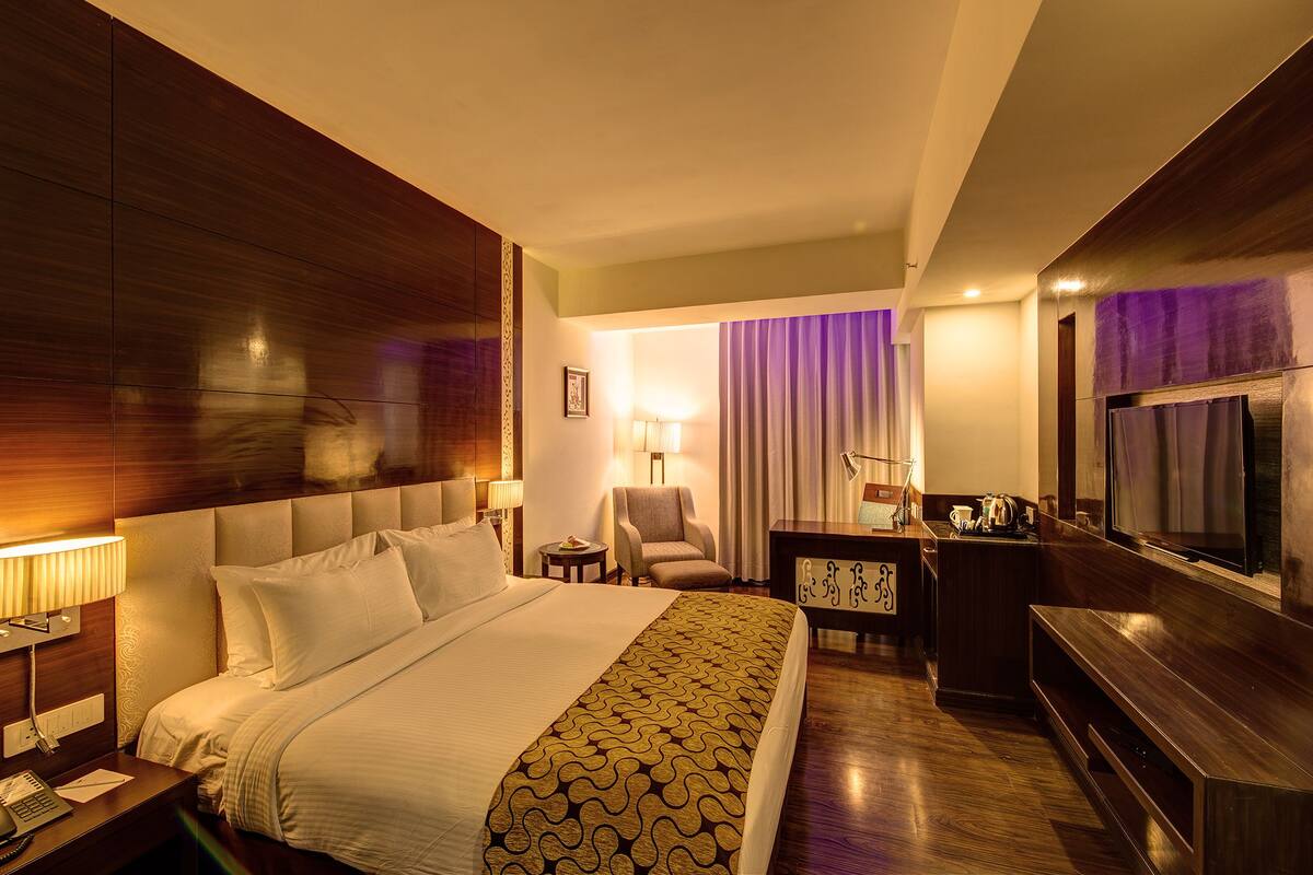Habitación de hotel con una cama grande, un escritorio con lámpara, una silla y un televisor.