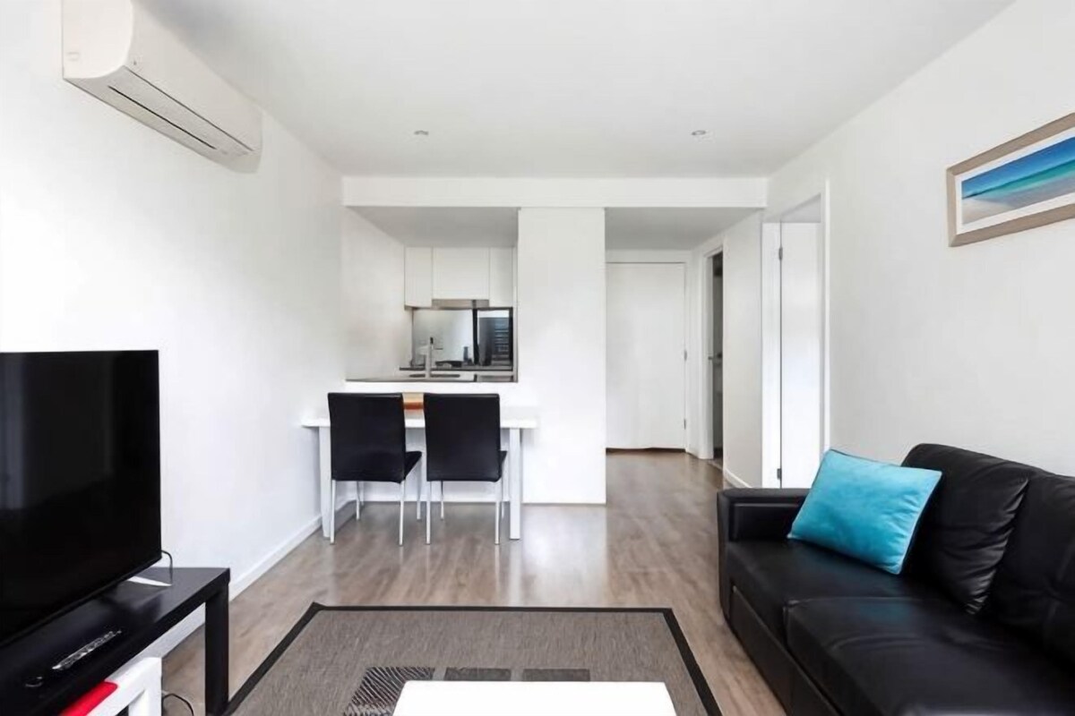 A modern living room with a flat-screen TV, a black leather sofa with a blue pillow, a dining area with a white table and chairs, and a kitchen in the background.