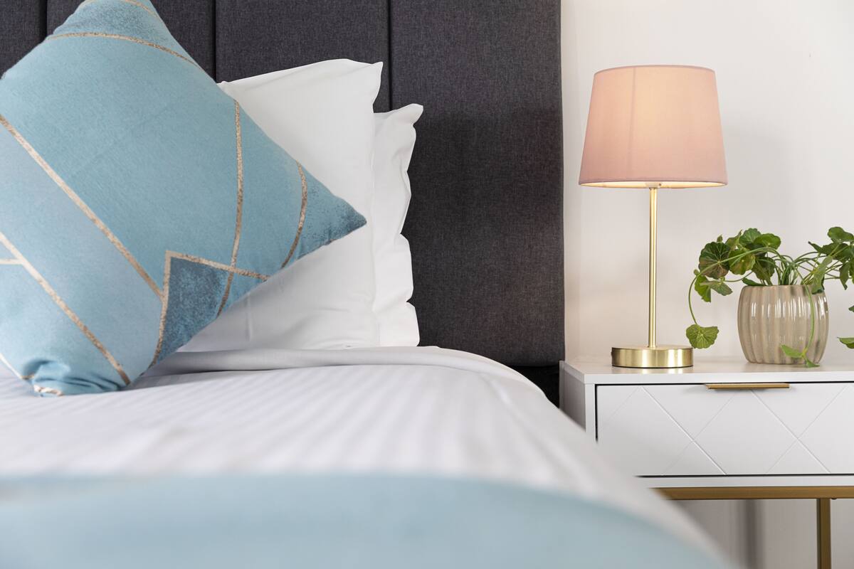 Ein ordentlich bezogenes Bett mit einem blau-weißen Kissen, einer Nachttischlampe und einem Topf mit einer Pflanze auf einem weißen Nachttisch.
