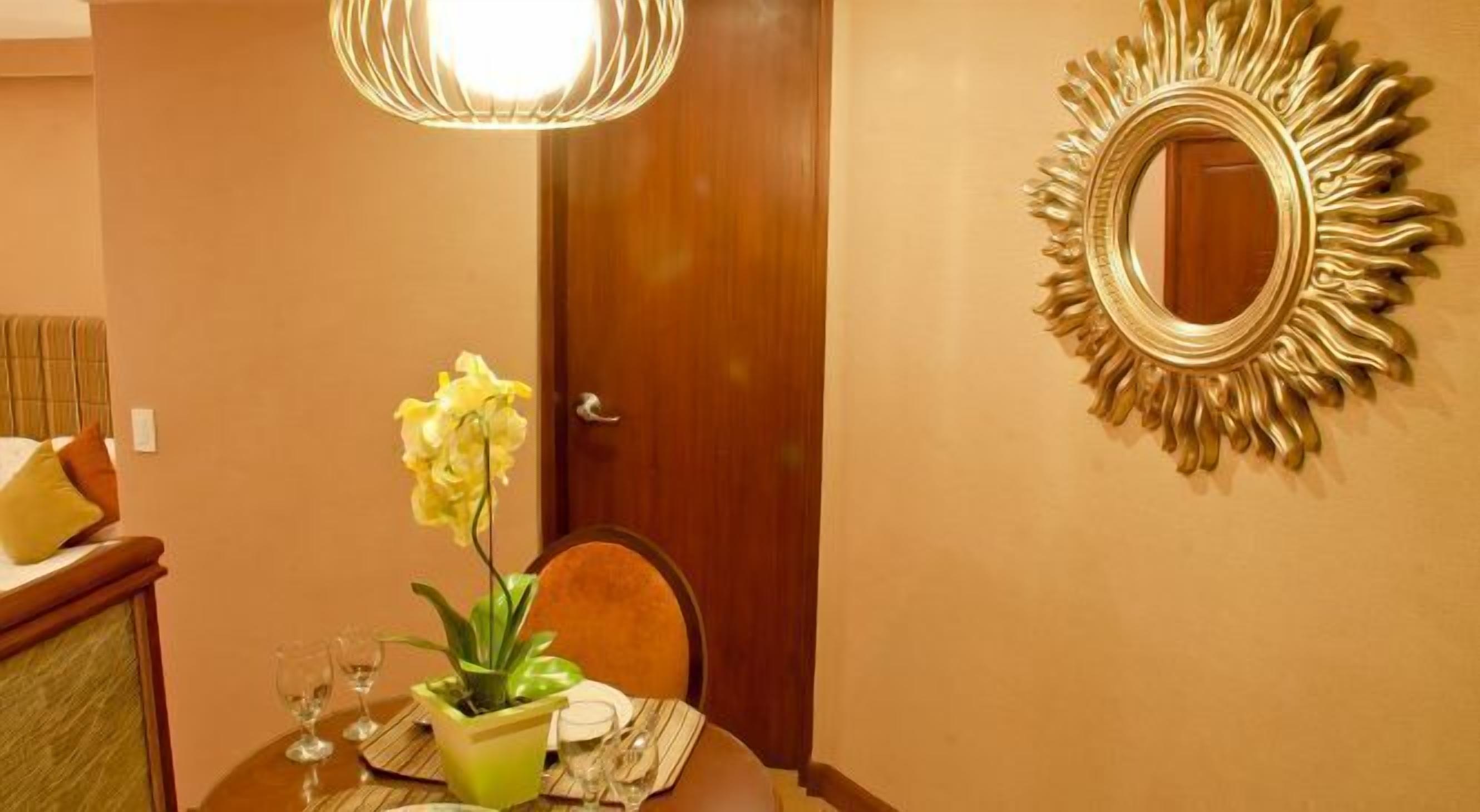 A dining area with a table set for two, a potted plant, and a mirror with a sunburst frame.