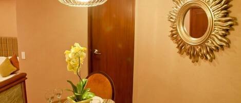 A dining area with a table set for two, a potted plant, and a mirror with a sunburst frame.