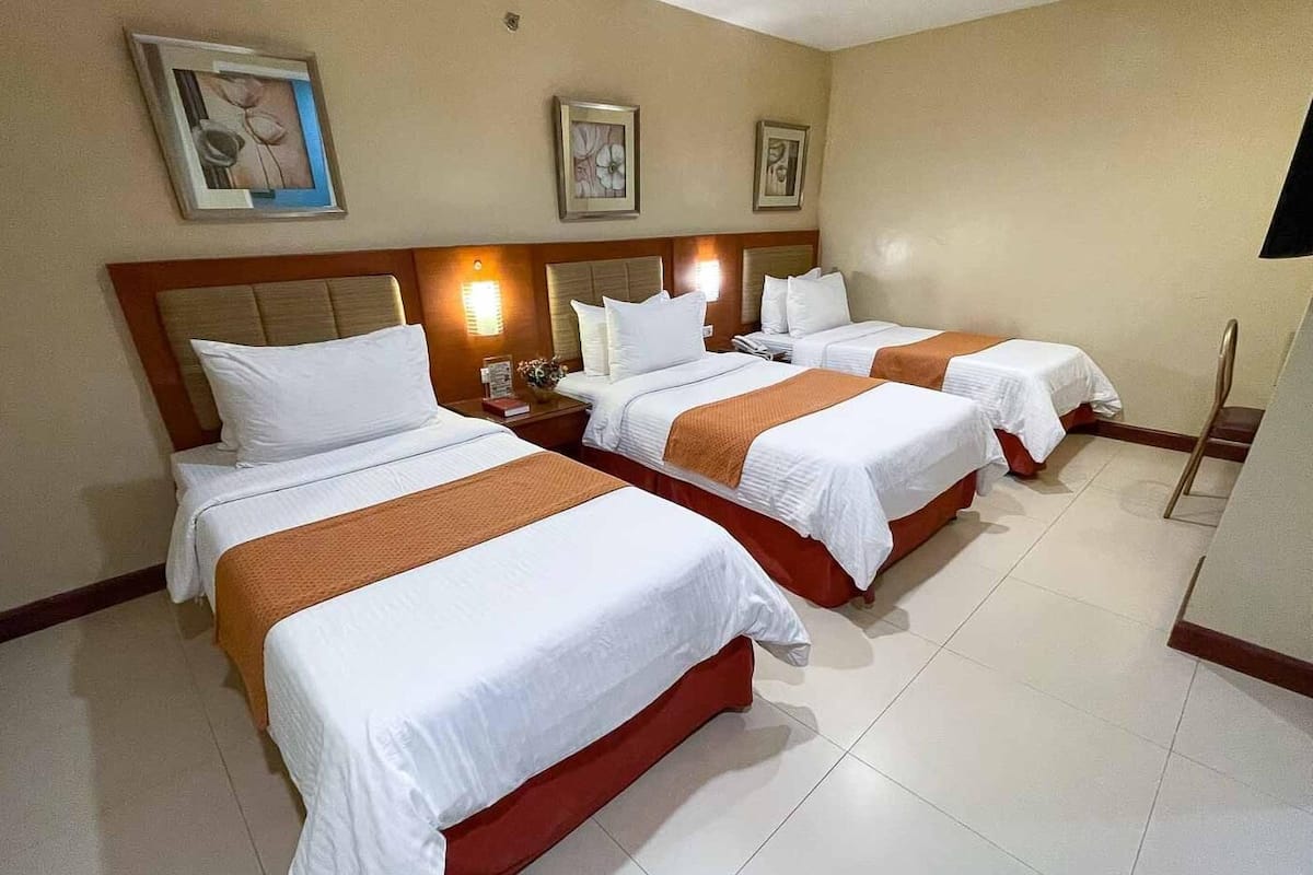 A hotel room with three beds, each with white linens and brown headboards. There are wall-mounted lights above each bed, and a flat-screen TV mounted on the wall to the right.