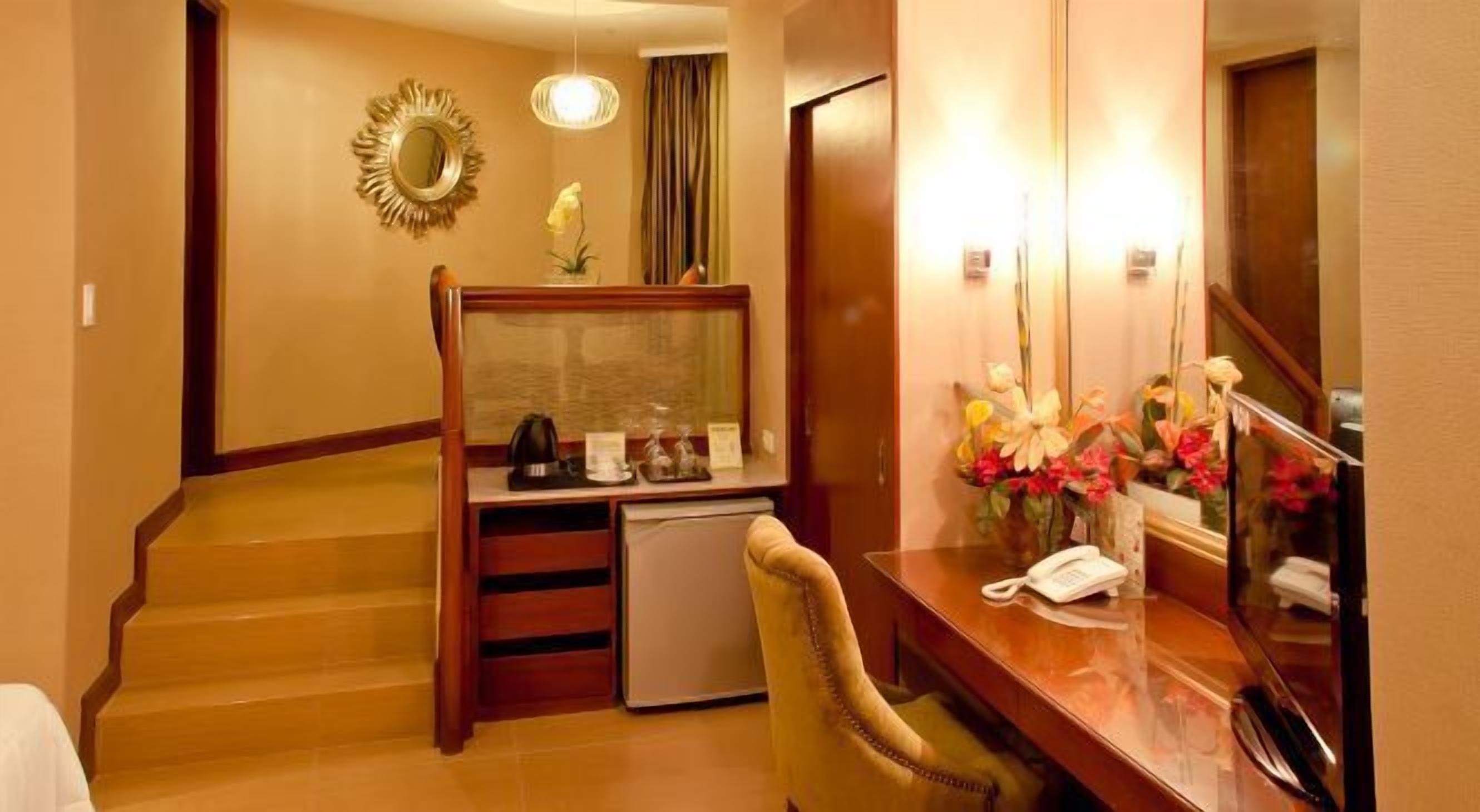 Hotelkamer met een houten bureau, telefoon, kleine koelkast en spiegel met bloemenarrangement.