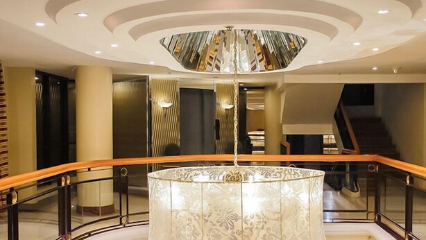 A modern hotel lobby with a central marble column, a wooden railing, and a unique ceiling design.