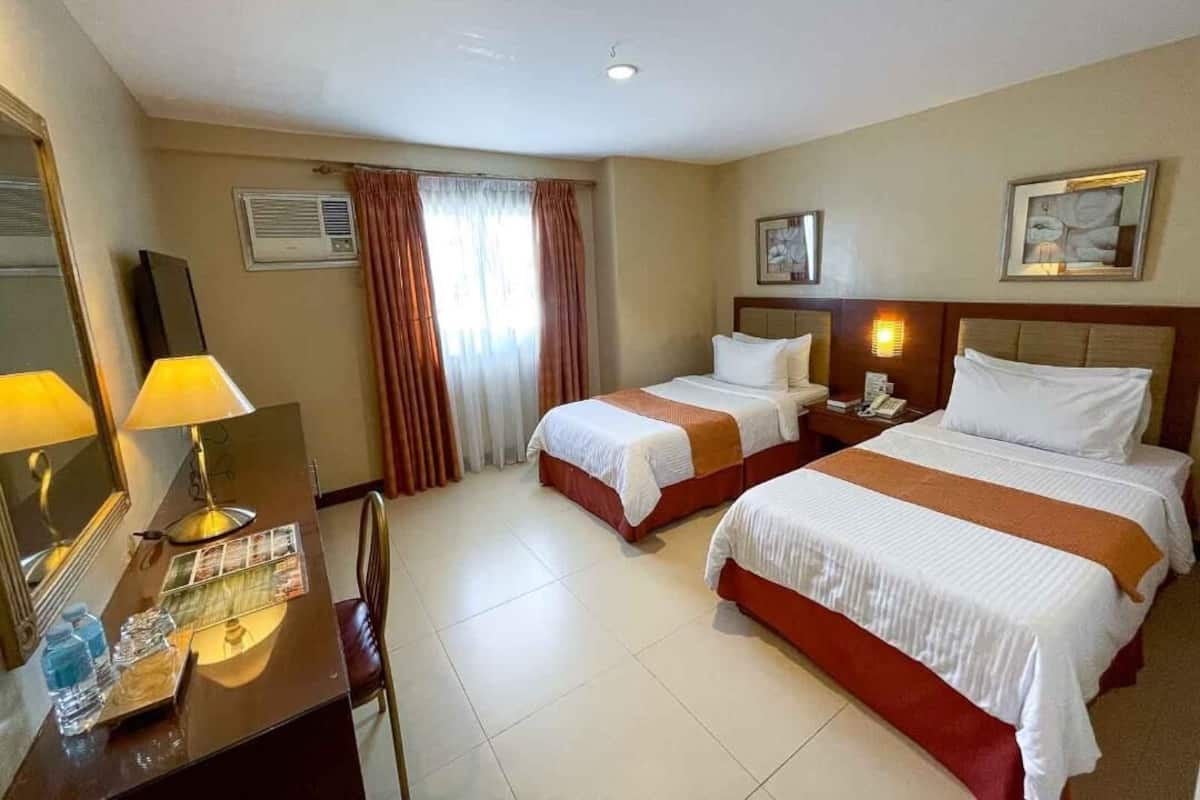 A hotel room with two beds, a desk with a lamp, a mirror, and a window with curtains.
