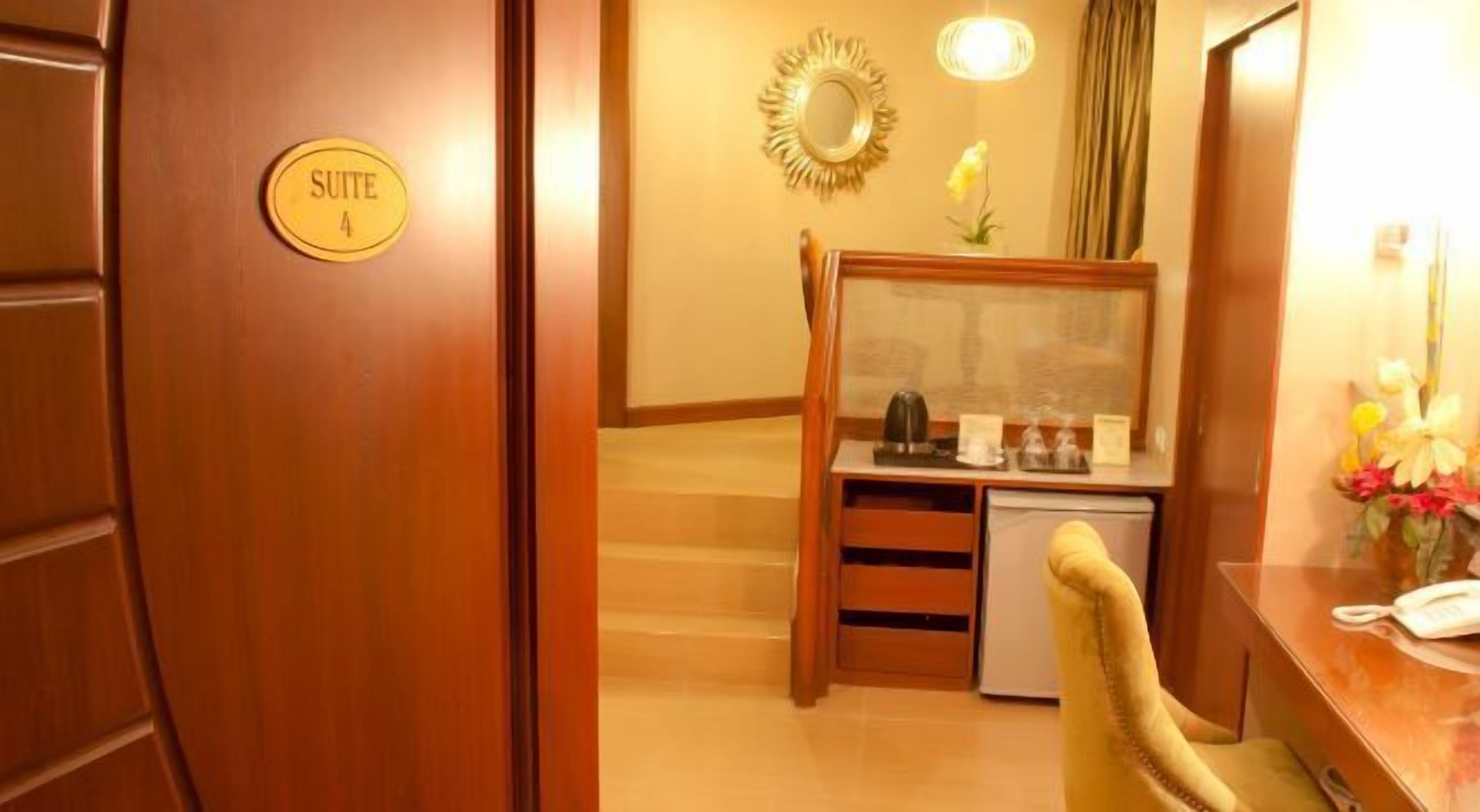 A hotel room with a wooden door labeled 'SUITE 4', a small desk with a telephone, a mirror, and a vase with flowers.