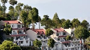 A cluster of white buildings with red roofs, surrounded by tall trees. - ECKO Woodsvilla Resort (Ranikhet)